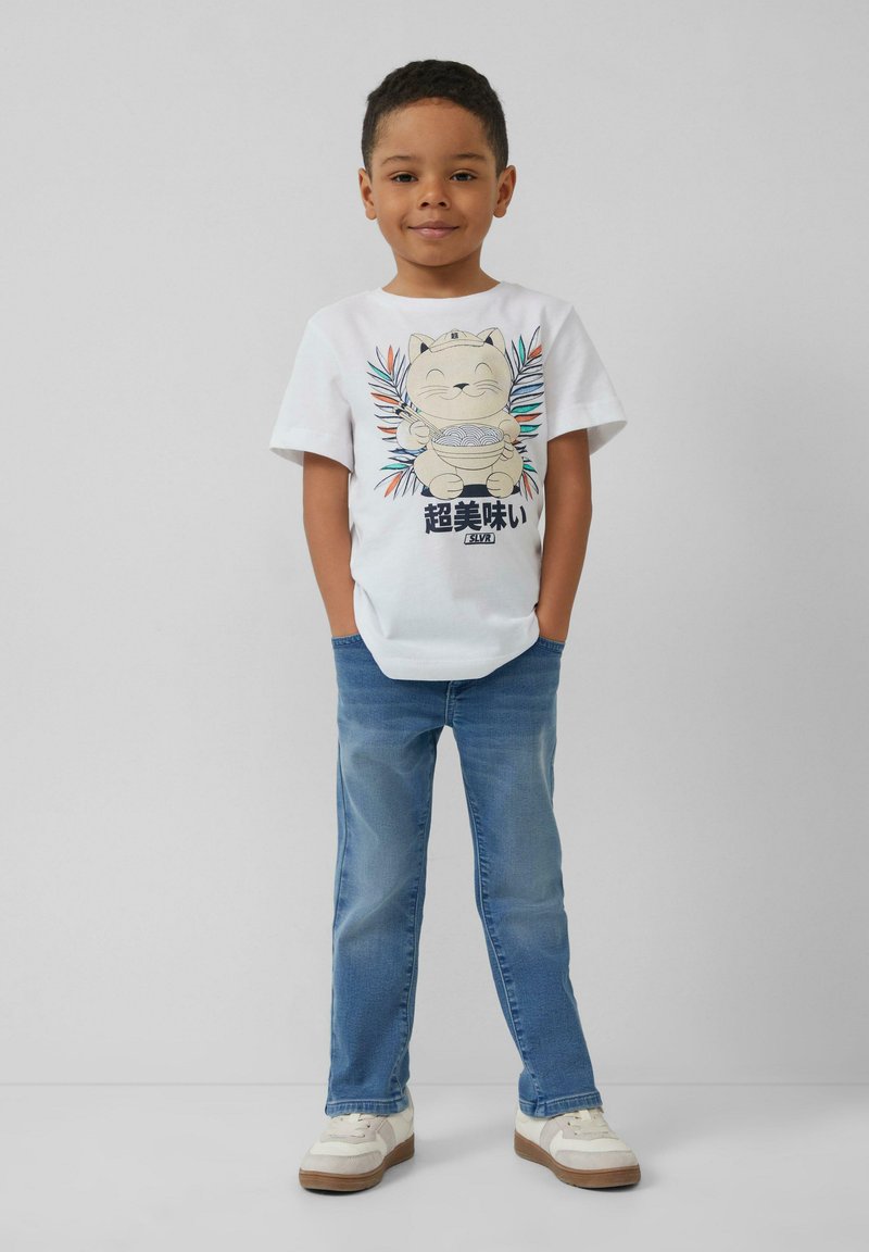 Witte katoenen t-shirt met een cartoon kat graphic, blauwe jeans en witte schoenen met beige accenten. De kat houdt noedels vast, omringd door kleurrijke bladeren.