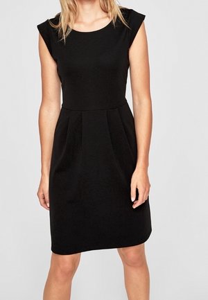 Žena v černých šatech bez rukávů, délky po kolena, s vypasovaným pasem a jemnými plisemi na jednoduchém pozadí.