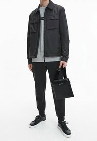 Giacca leggera nera con due tasche sul petto, felpa grigia, pantaloni neri e sneakers in pelle nera. Piccola borsa nera con tracolla.