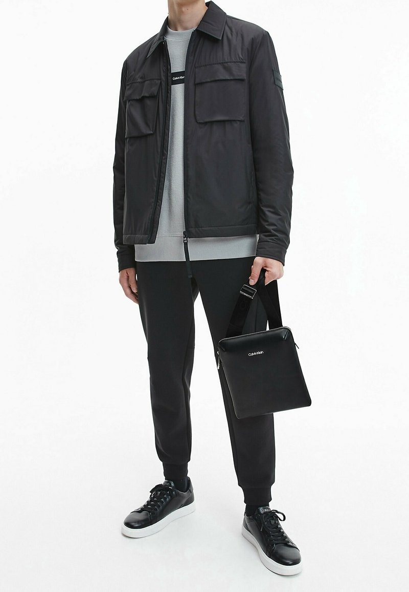 Giacca leggera nera con due tasche sul petto, felpa grigia, pantaloni neri e sneakers in pelle nera. Piccola borsa nera con tracolla.