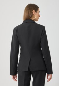 Μαύρο γάντζο κοστούμι με μακριά μανίκια, κολάρο notch και πίσω άνοιγμα. Λείο ύφασμα που προσφέρει μια δομημένη σιλουέτα για μια επίσημη εμφάνιση.