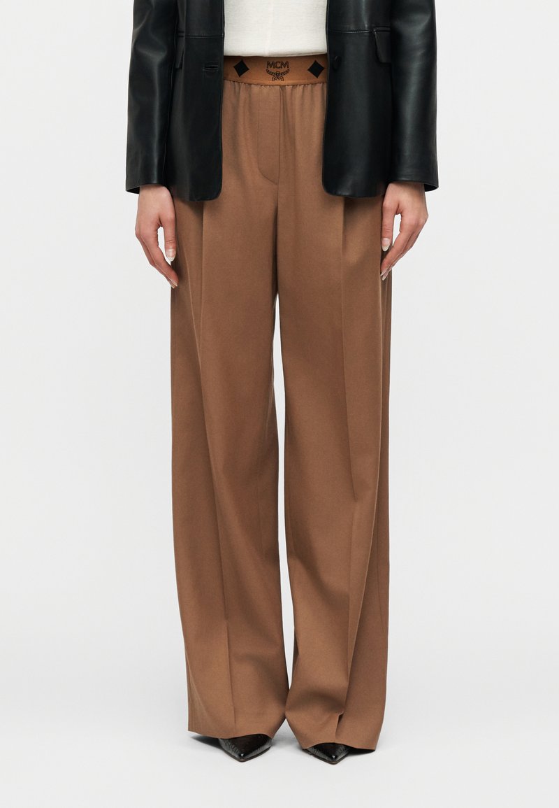 Personne portant un pantalon marron à taille haute et jambes larges, un blazer en cuir noir, et des chaussures noires pointues avec un logo visible sur la ceinture.