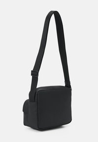 Czarna torba na ramię wykonana z gładkiego materiału syntetycznego. Ma kwadratowy kształt, z zamknięciem na zamek błyskawiczny oraz regulowanym paskiem ułatwiającym noszenie.