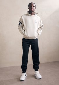 Beige hættetrøje med tre marineblå striber på ærmerne, parret med sorte bukser og hvide sneakers med sorte accenter. Glat tekstur.