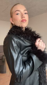 Zwarte leren jas met lange mouwen, voorzien van een zwarte bontkraag en een gestructureerde afwerking. Model houdt de jas dicht bij zich, waarmee het ontwerp wordt getoond.