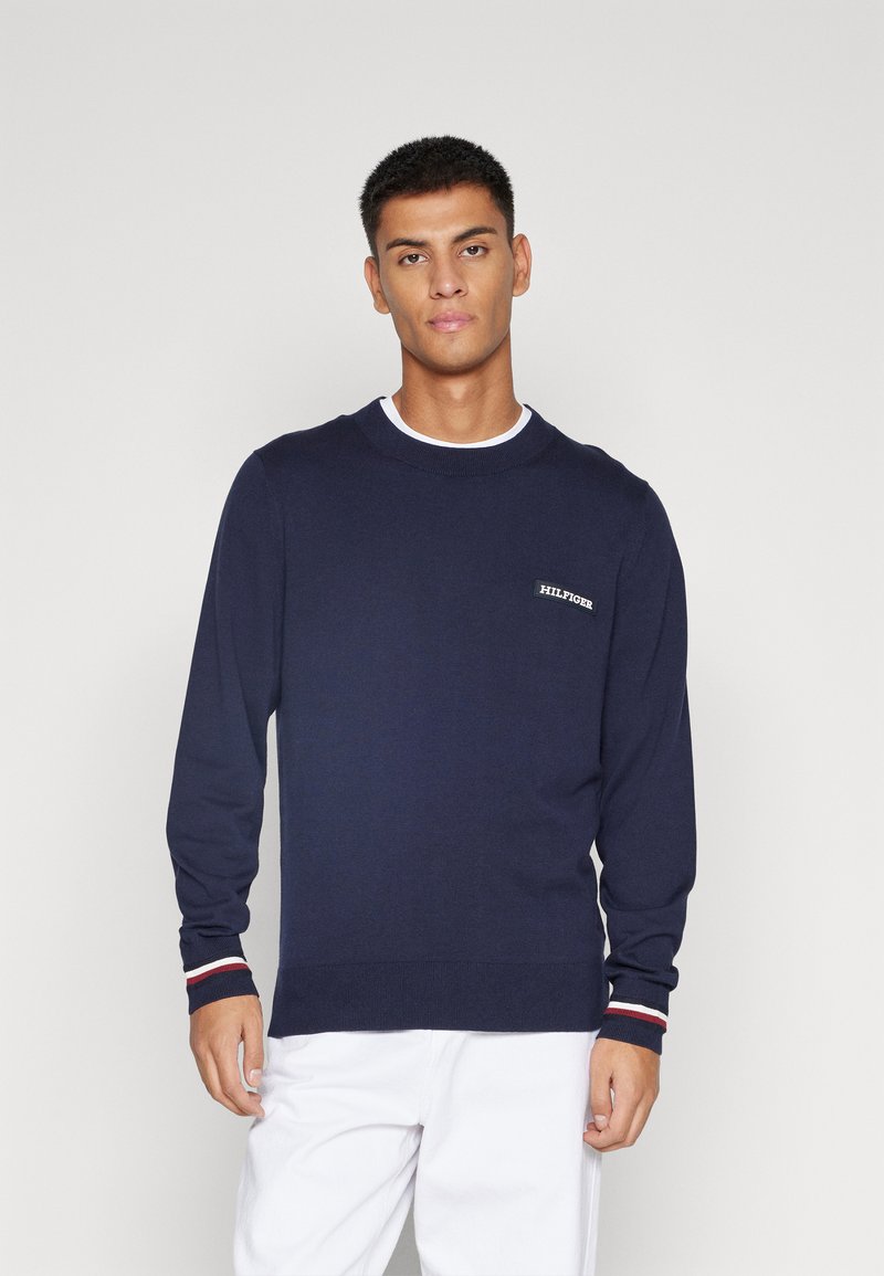 Maglione lavorato a maglia di colore blu navy con maniche lunghe, scollo rotondo e orlo a coste. Presenta un logo a contrasto sul petto e dettagli a righe sui polsini.