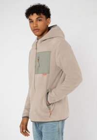 Beige Fleecejacke mit einer grünen Brusttasche, Reißverschluss und einer Kordelzugkapuze. Verfügt über orangefarbene Reißverschlussakzente und Seitentaschen.