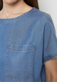 Top in denim azzurro chiaro con scollatura rotonda, maniche corte e una tasca applicata sul lato sinistro con dettagli di cucitura visibili.