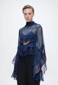 Γυναίκα που φορά διαφανές μπλε μπλούζα με βολάν στα φαρδιά μανίκια πάνω από ένα ταίριαστο τοπ, συνδυασμένη με φαρδιά μαύρη παντελόνα, στέκεται μπροστά σε λευκό φόντο.