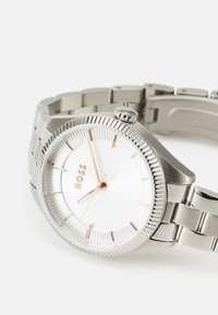 Sølvfarget rustfritt stålklokke med ribbet bezel, hvit urskive, rosegull timeindekser og en lenkearmbånd.