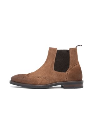Brauner Wildleder-Chelsea-Boot mit schwarzem elastischem Seiteneinsatz, Brogue-Details und niedrigem gestapeltem Absatz auf weißem Hintergrund.