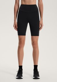 Fekete kerékpáros nadrág rugalmas anyagból, magas derékkal és sima tapintással, térd alatti hosszal, fekete sportcipővel párosítva.