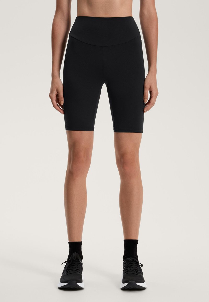 Fekete kerékpáros nadrág rugalmas anyagból, magas derékkal és sima tapintással, térd alatti hosszal, fekete sportcipővel párosítva.