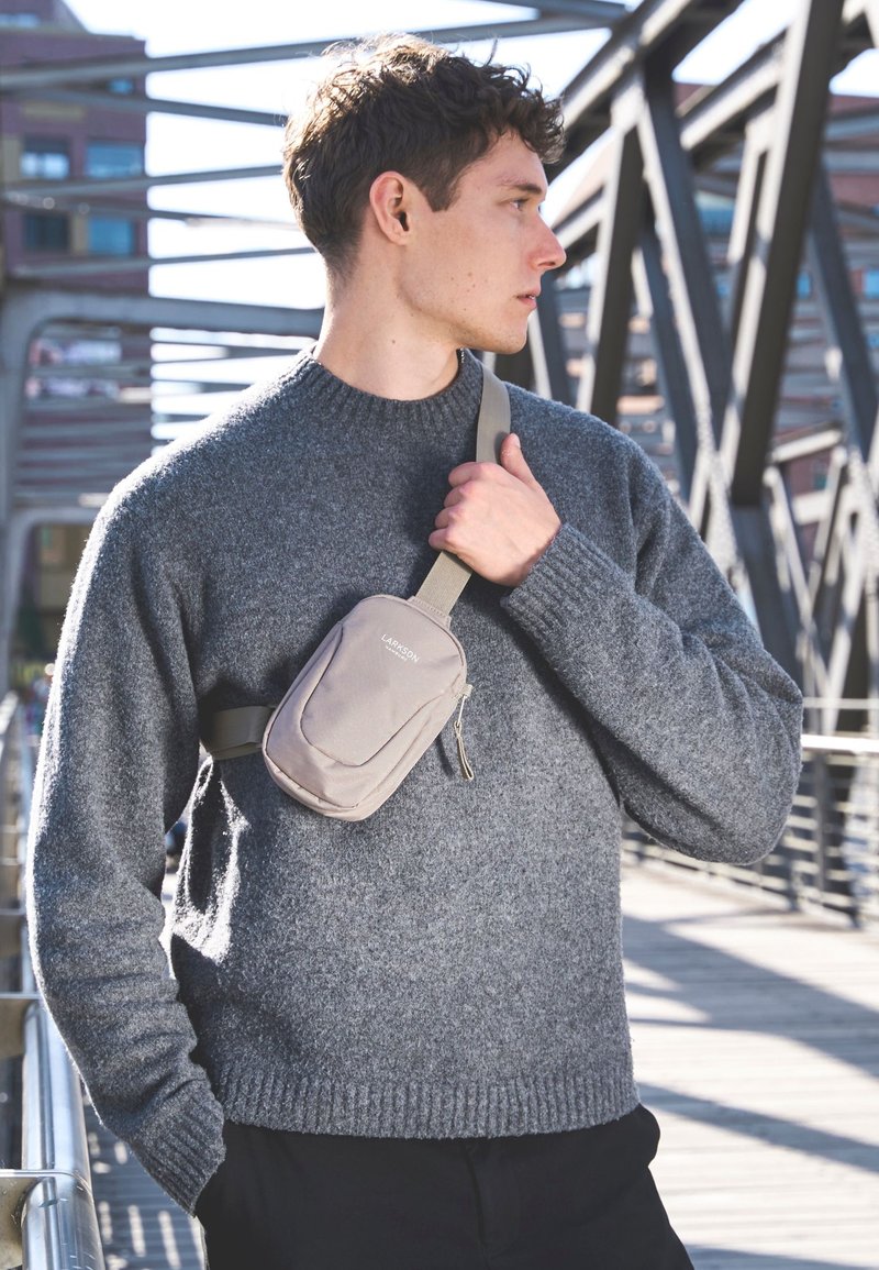 Maglione grigio lavorato a maglia con una finitura testurizzata, abbinato a una borsa a tracolla beige con tasca con zip e tracolla regolabile.