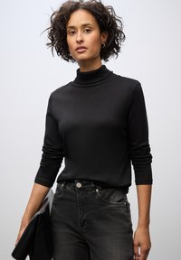 Maglione a collo alto nero con maniche lunghe, realizzato in un tessuto liscio. Abbinato a jeans neri. Il modello tiene una pochette nera. Sfondo semplice.