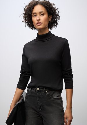 Sort turtleneck sweater med lange ærmer, lavet af et glat materiale. Kombineret med sorte jeans. Modellen holder en sort clutch. Ensfarvet baggrund.