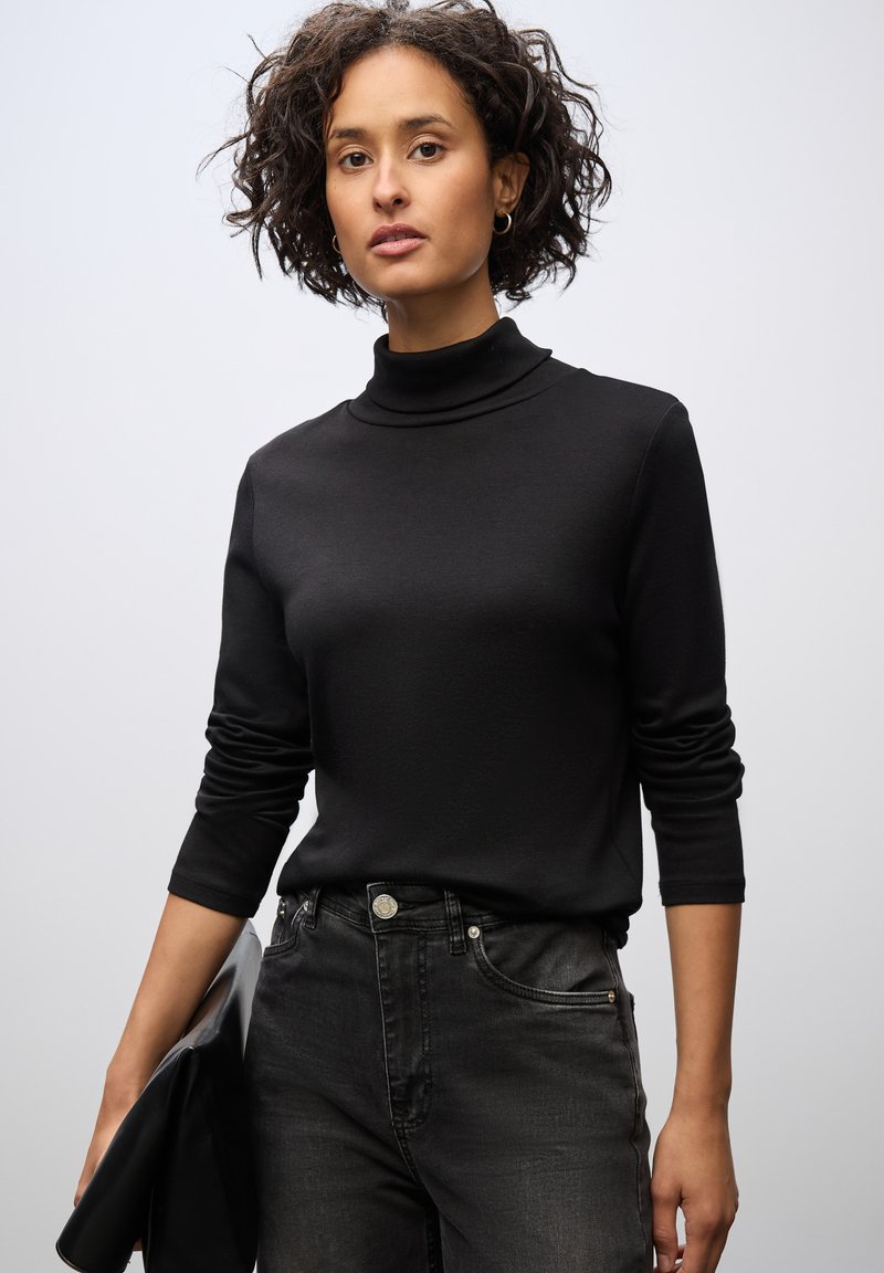 Maglione a collo alto nero con maniche lunghe, realizzato in un tessuto liscio. Abbinato a jeans neri. Il modello tiene una pochette nera. Sfondo semplice.