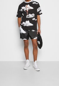 T-shirt graphique noir avec motif nuage, associé à un short noir présentant un motif similaire. Baskets blanches et sac Nike noir.