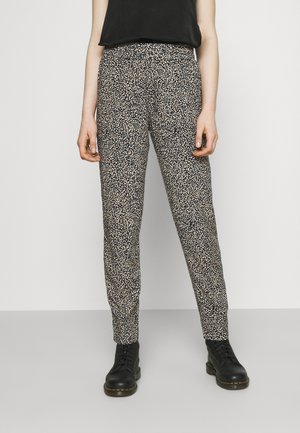 Mujer con pantalones negros con estampado floral, top negro sin mangas y botines negros con cordones, de pie sobre un fondo claro.