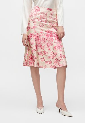 Femme portant une jupe droite crème avec imprimé floral rouge et architectural, un haut blanc à manches longues et des mules blanches à talons pointus.