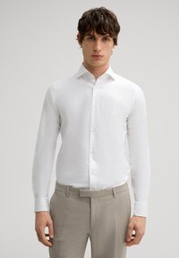 Camicia bianca a maniche lunghe con colletto alla francese, tessuto liscio e chiusura con bottoni, abbinata a pantaloni beige chiaro su misura.
