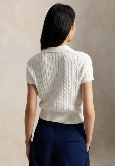 Woman with long dark hair wearing a white short-sleeve cable knit sweater and navy blue pants, facing away against a neutral background.