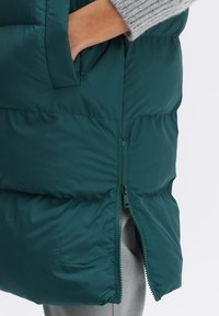 Türkiser Steppweste mit halblangem Reißverschluss, seitlichen Taschen und gestepptem Muster, kombiniert mit einem grauen Pullover darunter.