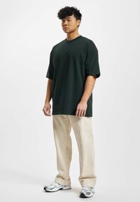 Tmavě zelené oversized tričko z bavlny, krátké rukávy, spárované s světle béžovými širokými kalhotami a sportovními bílými a zelenými teniskami.