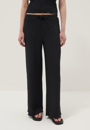 Pantalon noir texturé à jambes larges avec taille à cordon, porté avec des sandales noires à bout carré sur un fond clair.