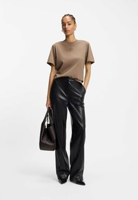 Donna che indossa una maglietta marrone aderente, pantaloni in pelle nera, scarpe nere a punta, mentre tiene in mano una borsa marrone scuro, in piedi contro uno sfondo bianco.