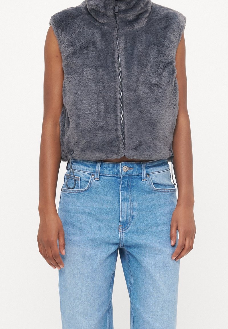 Gilet sans manches gris en fausse fourrure zippé, porté sur un jean taille haute bleu clair, sur une personne à la peau brune et aux bras détendus.
