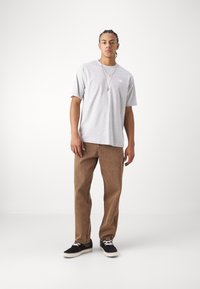 T-shirt gris à manches courtes avec un col ras du cou, associé à un pantalon marron à coupe droite. Baskets noires avec des accents blancs. Détail de collier subtil.