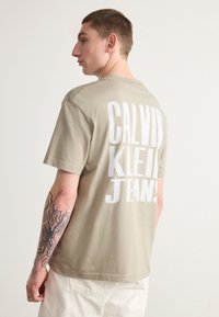 Bež pamukasta majica s velikim bijelim natpisom "CALVIN KLEIN JEANS" na leđima, kratkim rukavima i okruglim izrezom.
