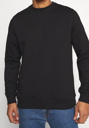 Homme portant un sweat-shirt noir uni à col rond et un jean bleu, debout sur un fond neutre.
