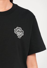 Μαύρη βαμβακερή μπλούζα με κοντό μανίκι, με άσπρο κεντητό λογότυπο ανθισμένου σήματος ειρήνης στο στήθος και κείμενο "SCH" και "JEANS" από κάτω.