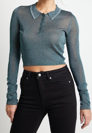 Vrouw draagt een glinsterende teal cropped top met lange mouwen en een kraag, gecombineerd met een zwarte jeans met hoge taille, tegen een effen achtergrond.
