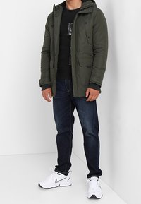 Olijfgroene, gewatteerde jas met capuchon en meerdere zakken aan de voorkant; gedragen over een zwarte gebreide trui, gecombineerd met donkere spijkerbroek en witte sneakers.