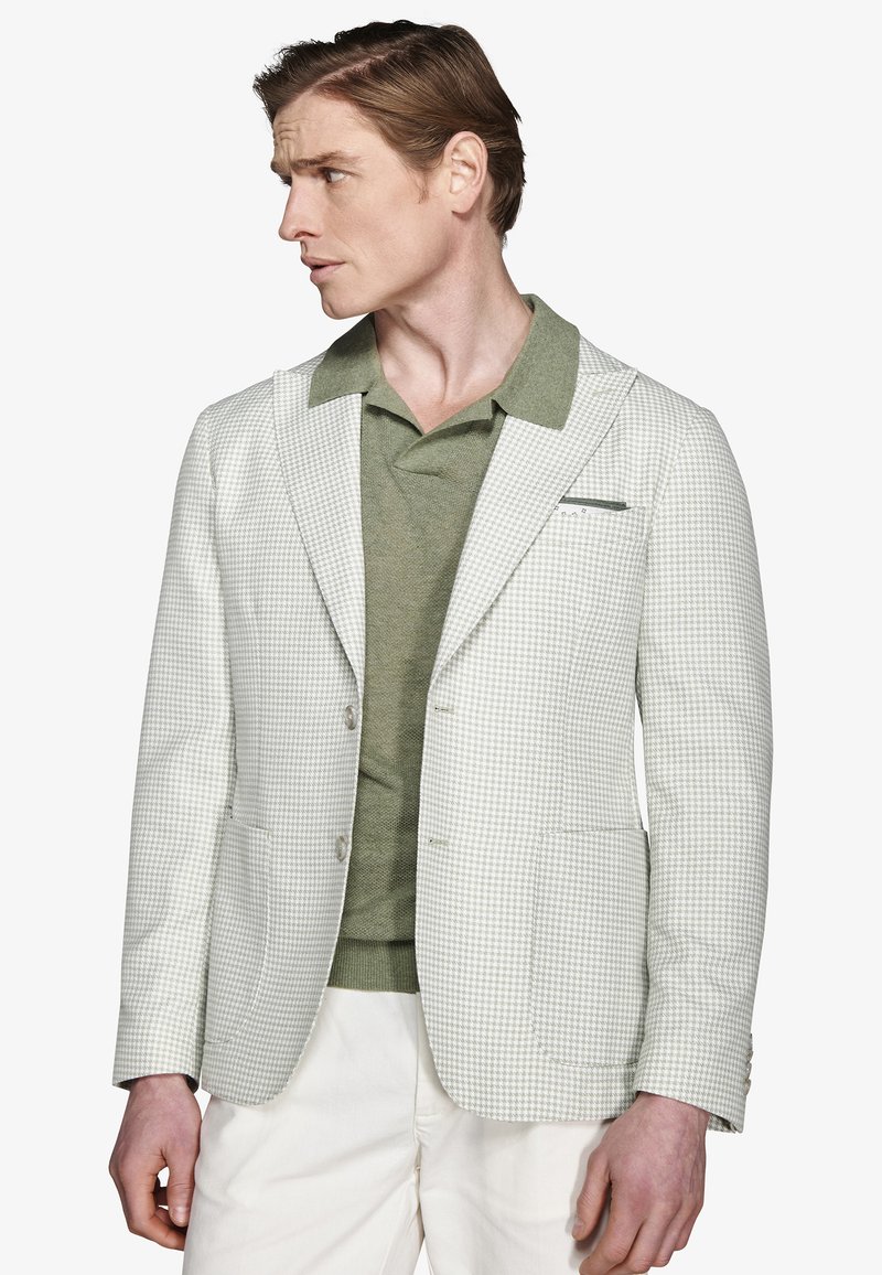 Hombre con blazer de cuadros gris claro sobre camisa polo verde y pantalones blancos, mirando hacia la izquierda contra un fondo blanco.