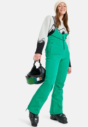 Femme portant un pantalon de ski vert, un pull à motif montagne, un bonnet en tricot blanc, tenant un casque de ski noir et des chaussures de ski noires.