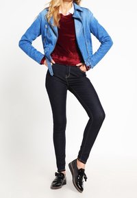 Chaqueta de moto de mezclilla en azul claro, combinada con una blusa de terciopelo burdeos y pantalones skinny oscuros. Zapatos de charol negros con detalle de lazo.