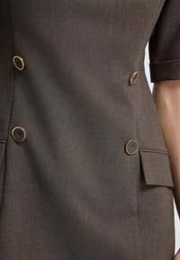 Hnědá přizpůsobená bunda se zlatými kulatými knoflíky, texturovaná látka, krátké rukávy a boční kapsy. Minimalistický design se strukturovaným tvarem.