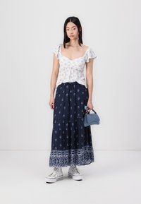Bluză albă cu flori și mâneci volanate, combinată cu o fustă maxi bleumarin cu modele albe și un tiv cu un model, și o geantă mică albastră.