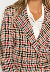 Dwurzędowa marynarka w wzór houndstooth, z czerwonymi, niebieskimi i czarnymi akcentami na beżowym tle. Widoczne detale w srebrnym kolorze.