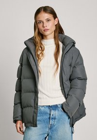 Pufová bunda v tmavě šedé barvě s polstrovaným designem, vysokým límcem a zipovým zapínáním. Na sobě má smetanový svetr s žebrováním a modré džíny.
