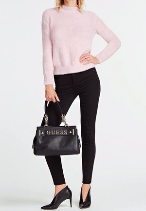 Woman wearing fluffy light pink sweater, black skinny pants, black pointed-toe heels, holding black GUESS handbag by handles.
