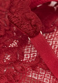 Czerwony koronkowy materiał z kwiatowymi wzorami, teksturowanym haftem oraz satynowym paskiem z małym metalowym pierścieniem.