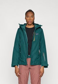 Teal waterdichte jas met een capuchon, voorzien van ritszakken en verstelbare manchetten, gedragen over een zwarte top en gecombineerd met roze broek.