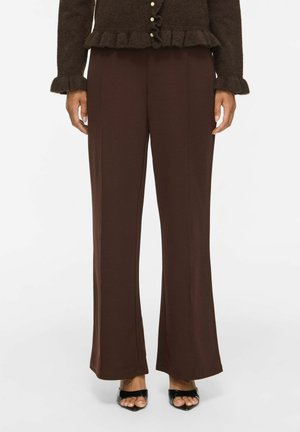 Femme portant un pantalon marron foncé à jambes larges, un pull marron texturé boutonné avec des bords volantés, et des chaussures à talons hauts noires ouvertes à l'avant.