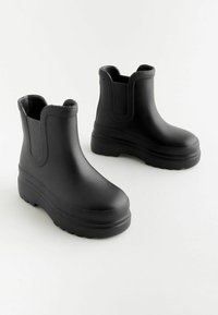 Bottines noires en caoutchouc avec semelles texturées, panneaux latéraux élastiques et finition lisse. Le design présente une pointe arrondie et une plateforme épaisse.