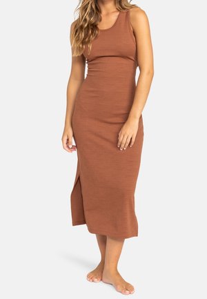 Vrouw draagt een mouwloze, nauwsluitende midi-jurk in roestkleur met een zijsplit, staat blootsvoets tegen een effen witte achtergrond.